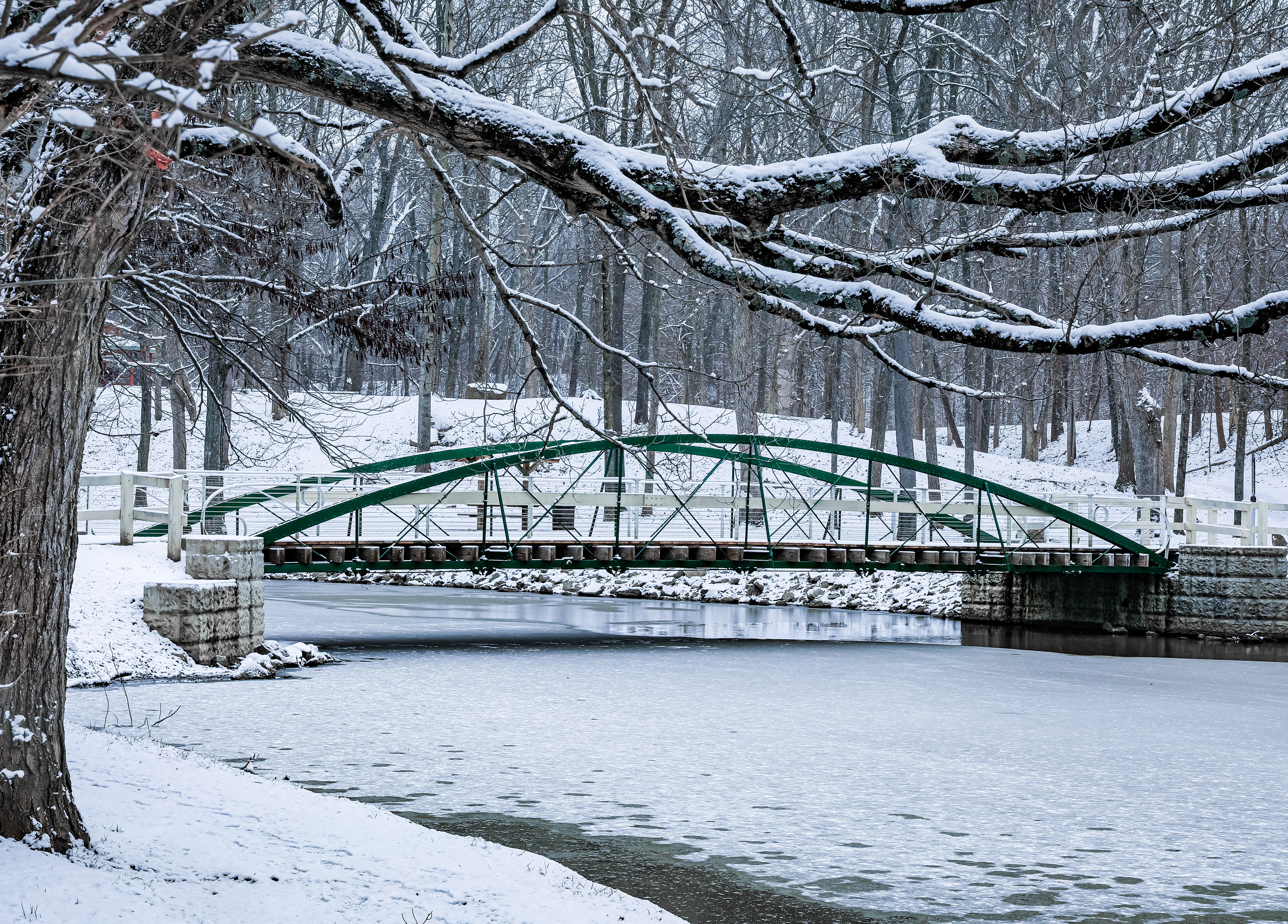 Historic green iron bridge crossing partially frozen water beneath snow-covered tree branches at Tawawa Park in Sidney Ohio